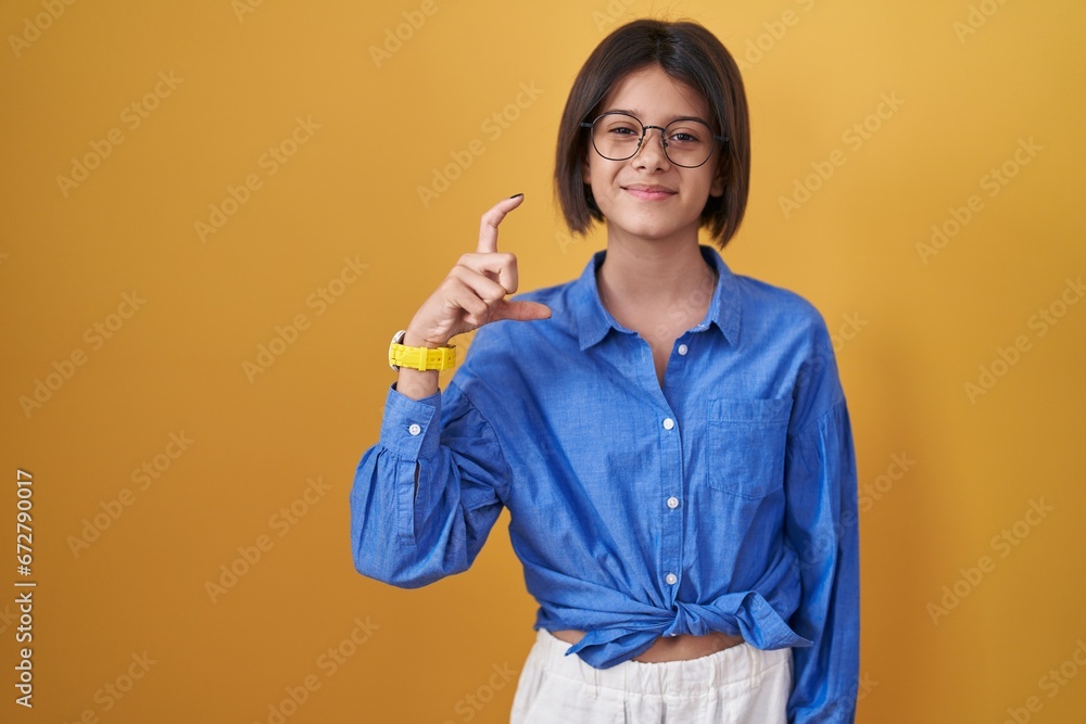 Young girl standing over yellow background smiling and confident gesturing with hand doing small size sign with fingers looking and the camera. measure concept. Young girl standing over yellow background smiling and confident gesturing with hand doing small size sign with fingers looking and the camera. measure concept.