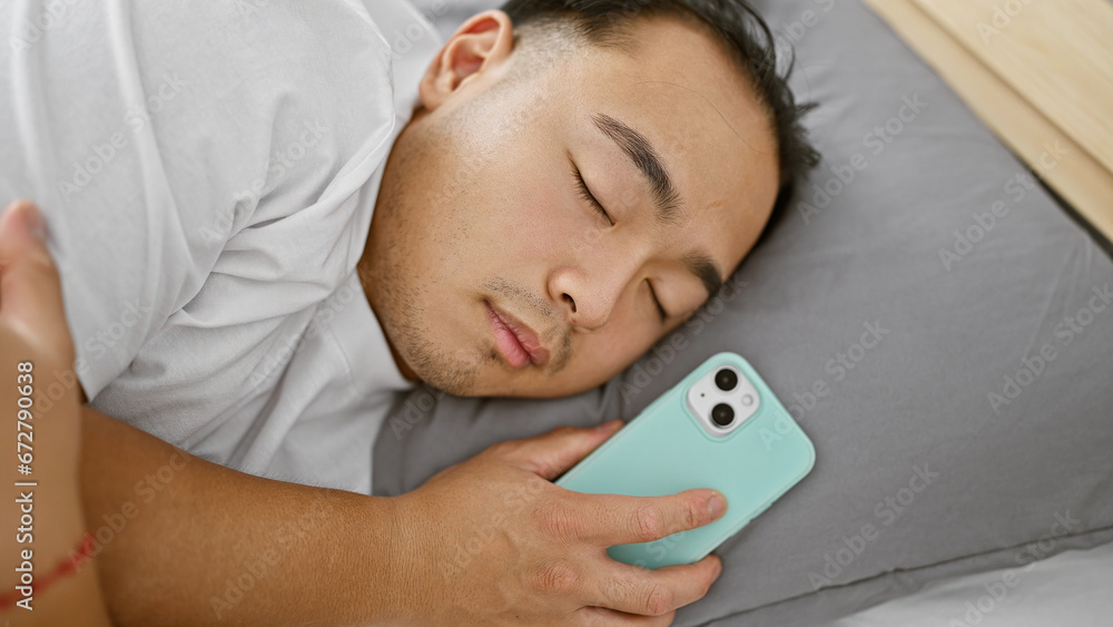 Exhausted young chinese man peacefully asleep in cozy bedroom, comfortably lying in bed with smartphone in hand, enjoying relaxed morning rest after a tiring day. Exhausted young chinese man peacefully asleep in cozy bedroom, comfortably lying in bed with smartphone in hand, enjoying relaxed morning rest after a tiring day.