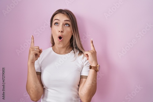 Blonde caucasian woman standing over pink background amazed and surprised looking up and pointing with fingers and raised arms. © Krakenimages.com