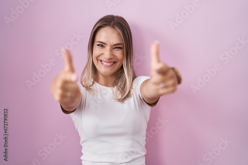 Blonde caucasian woman standing over pink background pointing fingers to camera with happy and funny face. good energy and vibes.