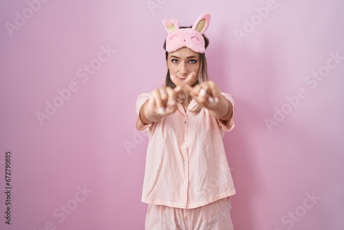 Blonde caucasian woman wearing sleep mask and pajama rejection expression crossing fingers doing negative sign