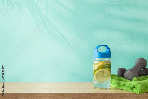 Fitness background with infused water bottle, towel and dumbbells on wooden table over blue background with palm tree shadows