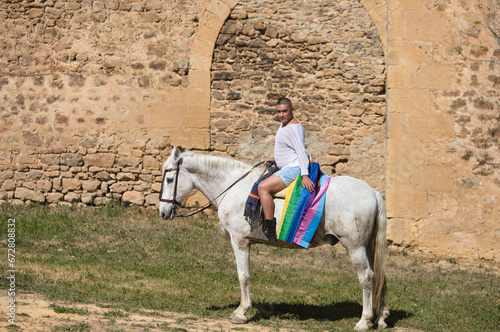 Young non-binary latin person, riding a white horse with the gay pride flag. In the background a medieval castle. Concept of diversity, homosexuality and human rights.