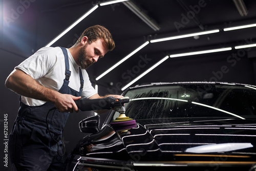 Side view on Young professional car service male worker with orbital polisher, polishing black luxury car hood in auto repair shop, copy space photo