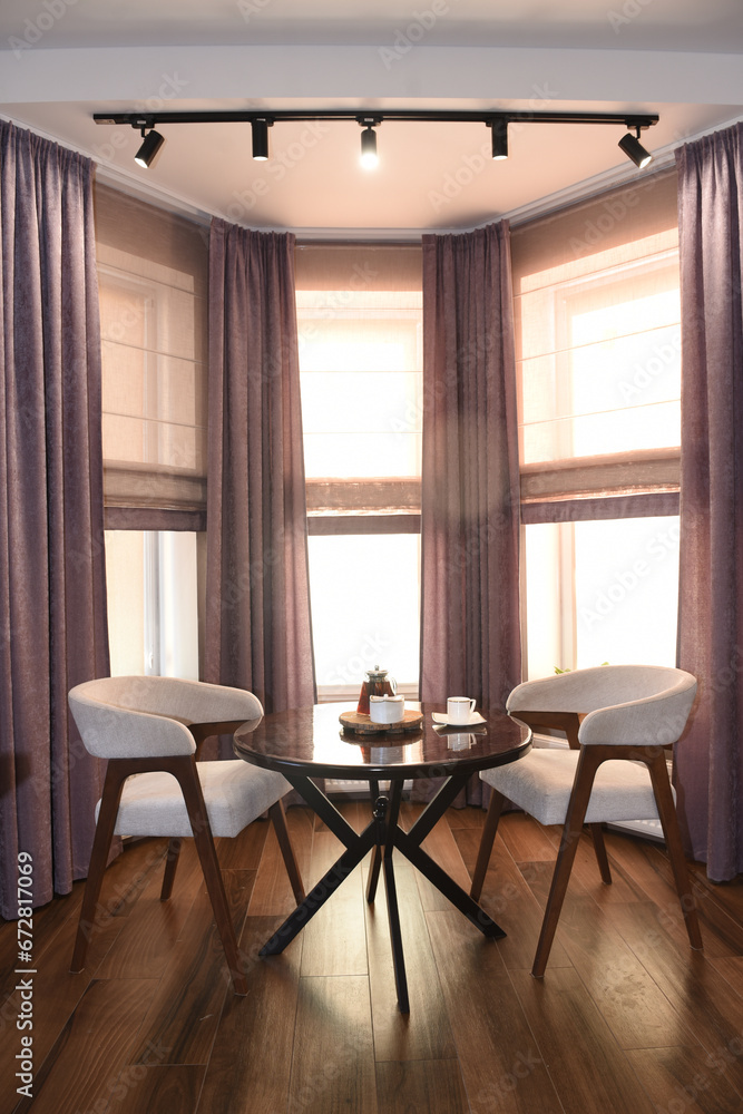 The room is cozy with curtains and a window with a table chairs and tea on the table