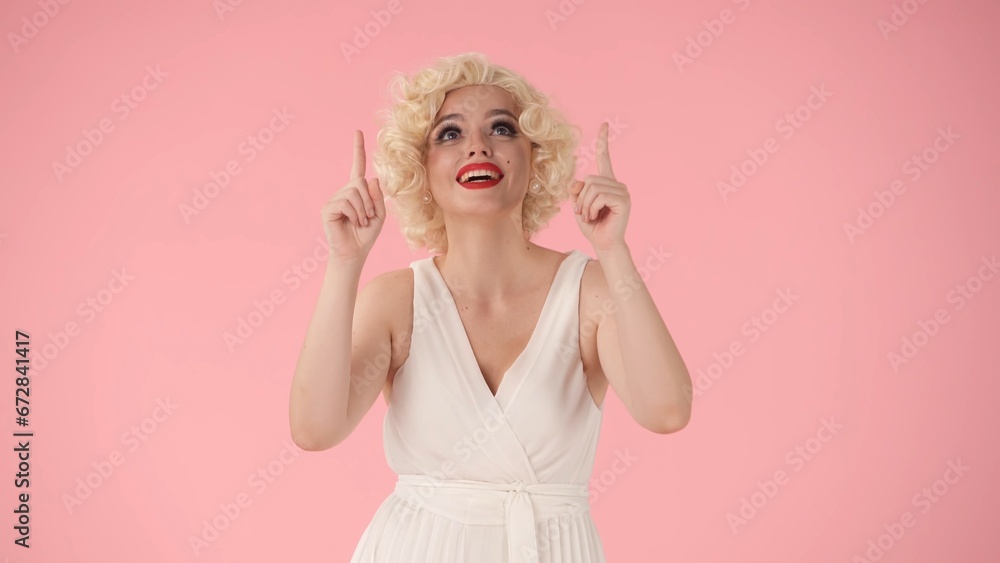 Woman points upward with her index fingers. Woman in white dress and wig, in the look of in studio on pink background. Advertising, promo, copy space. Woman points upward with her index fingers. Woman in white dress and wig, in the look of in studio on pink background. Advertising, promo, copy space.