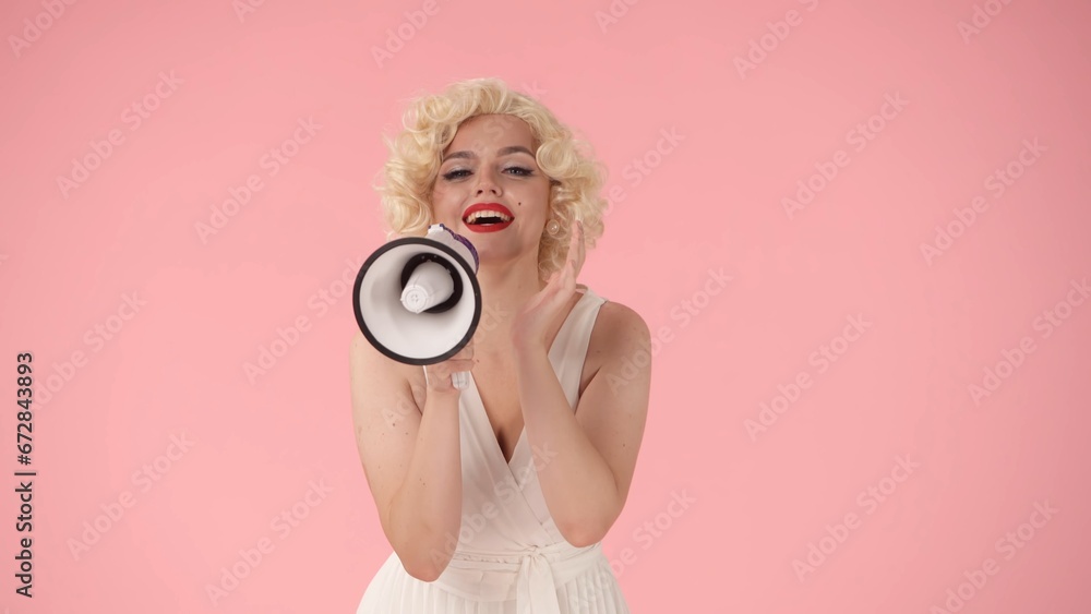 Obraz premium Woman in character of speaking into megaphone. Woman with colorful makeup, wig and white dress in studio on pink background. Sale, Black Friday. Copy space.