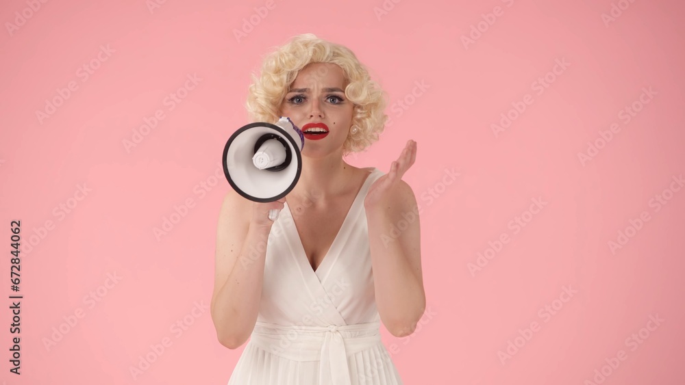Fototapeta premium Woman in character of speaking into megaphone. Woman with colorful makeup, wig and white dress in studio on pink background. Sale, Black Friday. Copy space.