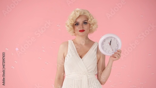 Woman holding a white round wall clock in her hand. Woman in image on , in studio surrounded by soap bubbles on pink background. Concept of time.