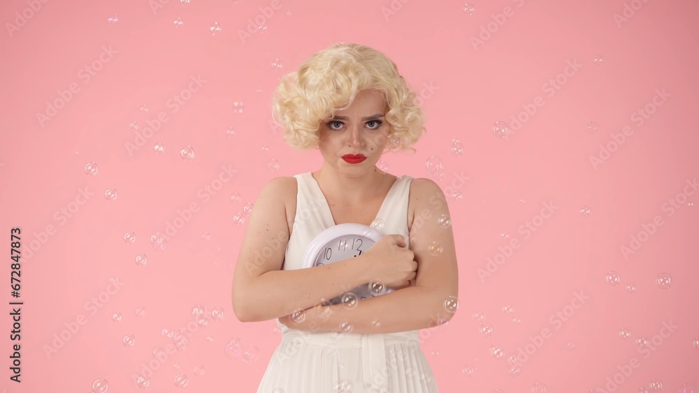Sad woman holding round clock, covering dial with hands. Woman in image on , in studio surrounded by soap bubbles on pink background. Concept of time. Sad woman holding round clock, covering dial with hands. Woman in image on , in studio surrounded by soap bubbles on pink background. Concept of time.