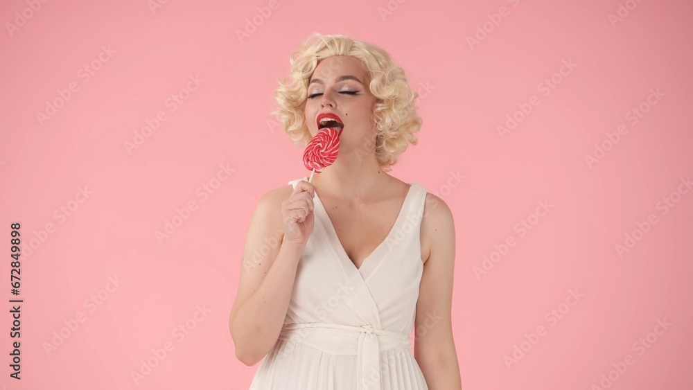 Woman happily licking a colorful lollipop on a stick. Woman in the image of in studio on pink background. Sweet food concept. Woman happily licking a colorful lollipop on a stick. Woman in the image of in studio on pink background. Sweet food concept.