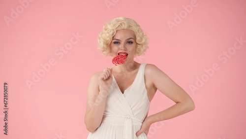 A female coquette bites a colorful lollipop with her teeth. Woman in the image of in studio on pink background. Sweet food concept.