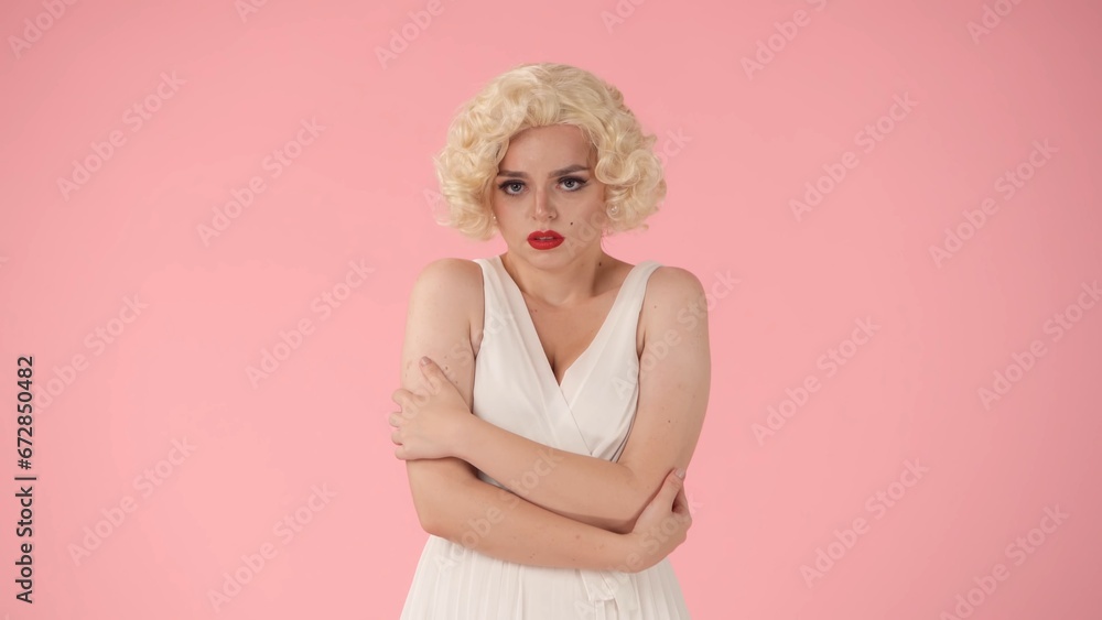 A young woman stands offended. Portrait of a woman in the image of , wearing a white dress and white wig in a studio on a pink background. A young woman stands offended. Portrait of a woman in the image of , wearing a white dress and white wig in a studio on a pink background.