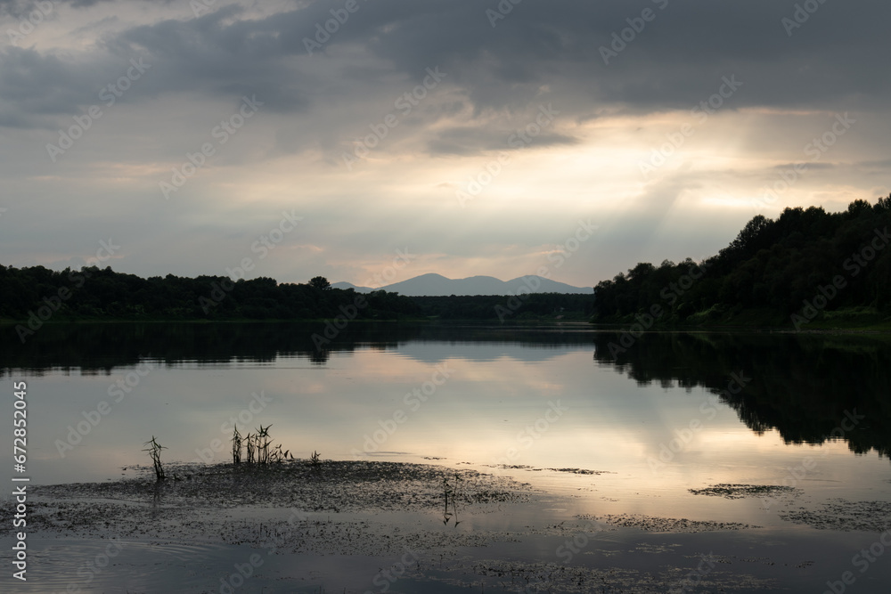 Landscape with mountain silhouette reflecting in river and overcast clouds with glowing sun rays