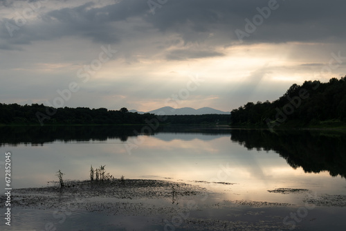 Landscape with mountain silhouette reflecting in river and overcast clouds with glowing sun rays