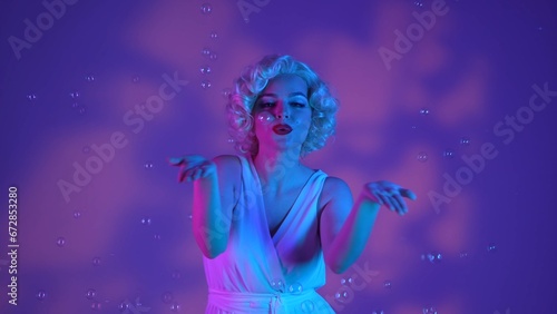 Woman dancing surrounded by soap bubbles, sending air kisses. Woman in the image of on a pink background with highlights, in a studio illuminated with pink and green neon light.