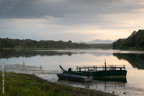 Landscape with moored boats in river against mountain silhouette in distance and cloudy sky