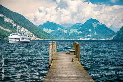 A ship cruises on Lake Lucerne with rippled waters, featuring a pier in the foreground. Enjoy the cloudy skies, deep blue waters, and lush green Alpine hills. Scenic Swiss Mountain Landscape. © Werner