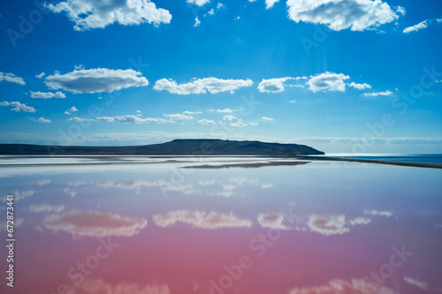 A picturesque pink lake under a blue cloudy sky with a mirror reflection of clouds in the water. In the distance are white salt deposits and the seashore. Filming from a drone.