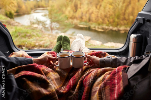 travel, tourism and camping concept - view to river from car trunk with couple under blanket toasting tea cups