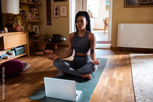 Young woman meditating with laptop on mat at home photo
