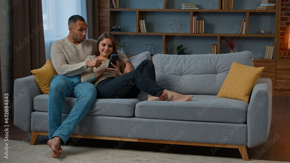 Diverse man and woman interracial couple homeowners relax on sofa talk video call communicate online with mobile phone web app happy diverse wife husband buy internet goods chatting read social media Diverse man and woman interracial couple homeowners relax on sofa talk video call communicate online with mobile phone web app happy diverse wife husband buy internet goods chatting read social media