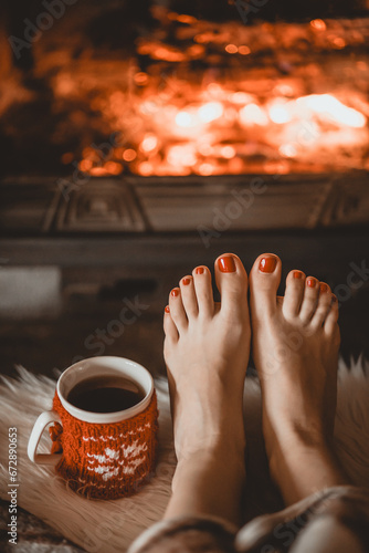 Bare woman feet by the cozy fireplace. Woman relaxes by warm fire with a cup of hot drink and warming up her feet. Close up on feet. Winter and Christmas holidays concept.