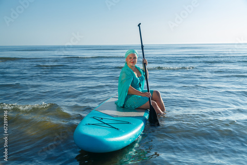 A woman in a turquoise swimsuit with a skirt and a scarf on her head sits on a SUP board near the sea