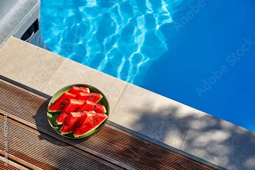 A bright red cut watermelon lies near the swimming pool. High angle. Copy space. Blue water by the pool. Holidays in resorts and travel.