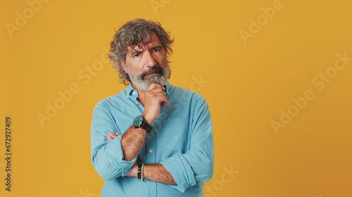 Elderly gray-haired bearded man wears a blue shirt, looks at the camera listens carefully and agrees, isolated on an orange background in the studio