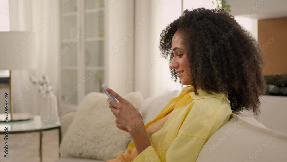 Smiling African American girl woman play mobile video game on home couch in living room happy relaxed ethnic female teenager scrolling smartphone social networks addict internet dating apps side view Smiling African American girl woman play mobile video game on home couch in living room happy relaxed ethnic female teenager scrolling smartphone social networks addict internet dating apps side view
