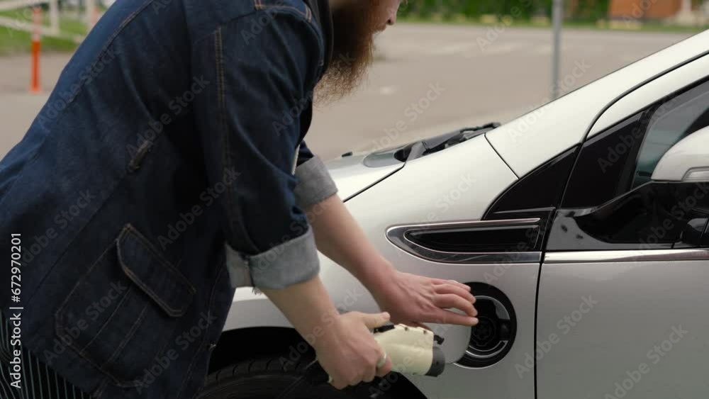 Bearded man inserts power connector into an electric car. Guy in denim jacket connects charging cable to an electric car and charges the batteries. Electric car station, zero emissions Bearded man inserts power connector into an electric car. Guy in denim jacket connects charging cable to an electric car and charges the batteries. Electric car station, zero emissions