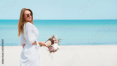 Joyful Young Woman Holding a Bouquet of Flowers on a Beach with Blue Sky Background