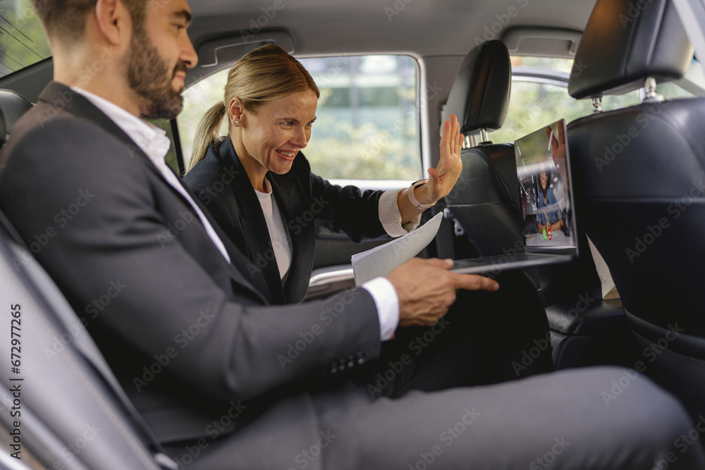 Smiling business colleagues have online meeting using laptop while sitting in car back seats Smiling business colleagues have online meeting using laptop while sitting in car back seats