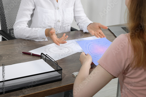 Astrologer showing zodiac wheel to client at wooden table indoors, closeup