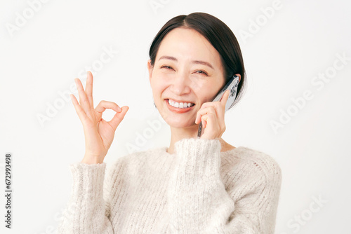 スマートフォンで通話しながらOKサインをする女性 白背景