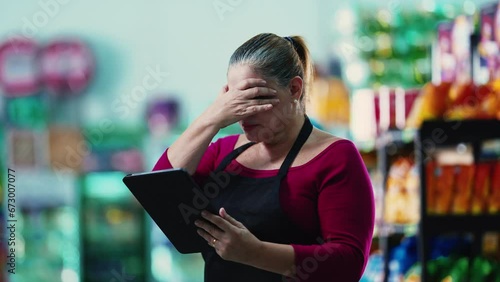 Stressed female entrepreneur owner of small business struggling with hard times holding tablet and staring at the bottom line of the grocery store photo