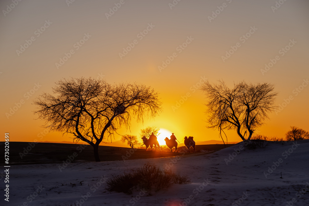 Silhouette of a Mongolian riding a camel in the snow-covered desert as the sun sets.