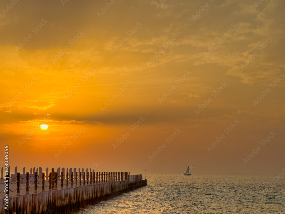 Sunset at a pier with a small sailboat in the background.