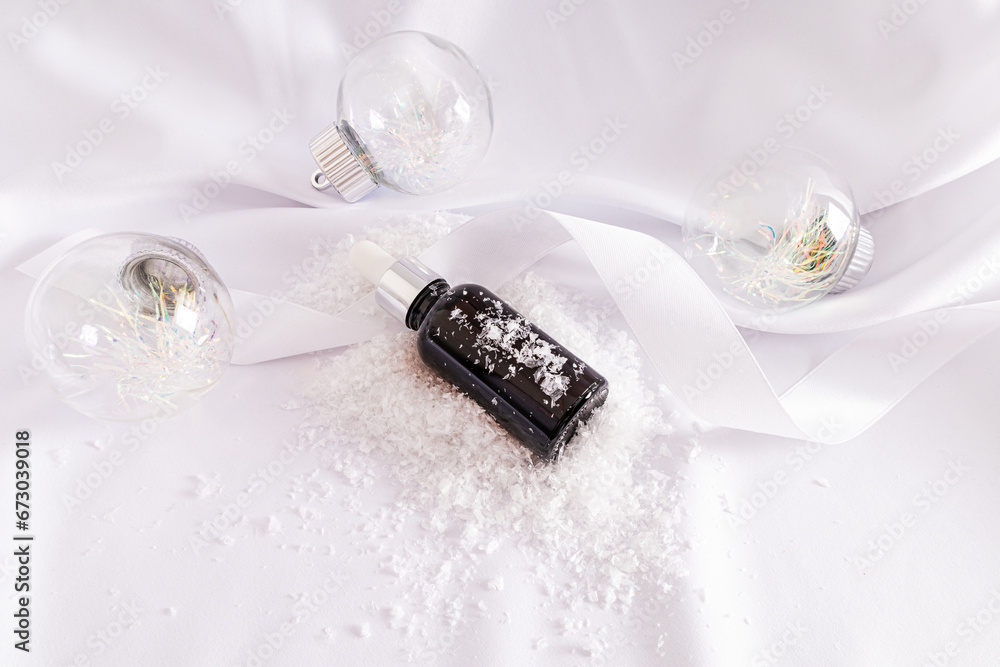 Glass cosmetic bottle with dropper lies on white satin and artificial snow among fashionable glass balls. New Year cosmetics concept. Glass cosmetic bottle with dropper lies on white satin and artificial snow among fashionable glass balls. New Year cosmetics concept.