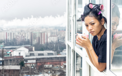 A young woman in curlers looks out of the window of a high-rise building in an industrial zone photo