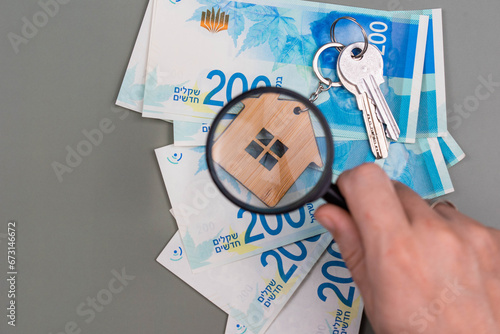 Buying a house, building repair and mortgage concept in Israel. Estimation real estate property with loan money and banking. Keys and house with shekel cash banknotes under magnifying glass. photo