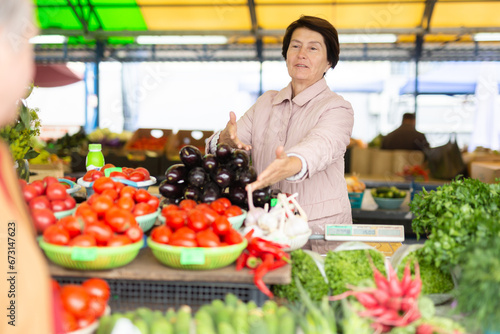 Smiling elderly female seller telling prices for fresh vegetables at local bazaar