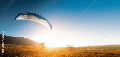 A paraglider glides along the ground at sunset with mountains in the background. Panoramic shot banner for paragliding in warm colors. Glare from the sun in the frame