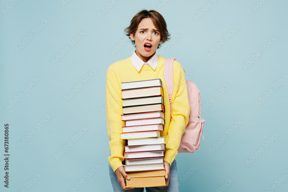 Young shocked dissatisfied indignant woman student wear casual clothes yellow sweater backpack bag hold stack of many books isolated on plain blue background. High school university college concept. Young shocked dissatisfied indignant woman student wear casual clothes yellow sweater backpack bag hold stack of many books isolated on plain blue background. High school university college concept.