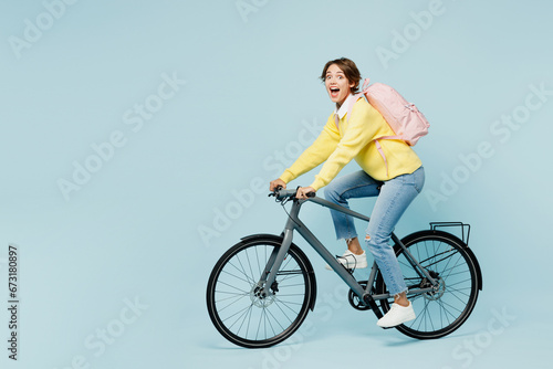 Full body side view young shocked surprised woman student wears casual clothes sweater backpack bag ride bicycle look camera isolated on plain blue background. High school university college concept.