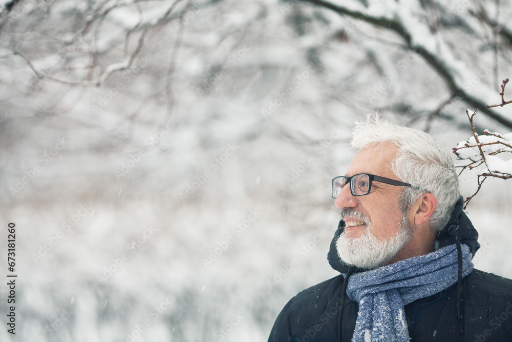 Active grandpa concept. Portrait of handsome mature man in trendy winter clothing and fashionable glasses standing under tree, over snow park background. Close up. Text space.  Outdoor shot