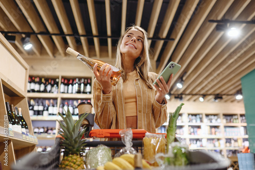 Young customer woman wear casual clothes hold wine use mobile cell phone shopping at supermaket store grocery shop buying with trolley cart choose products inside hypermarket. Purchasing food concept.