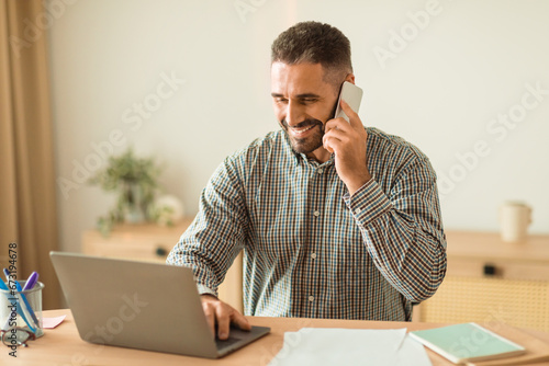 Cheerful Businessman Talking On Phone At Laptop In Office Interior