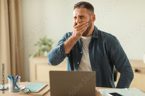 Tired man yawning while sitting at laptop in modern office